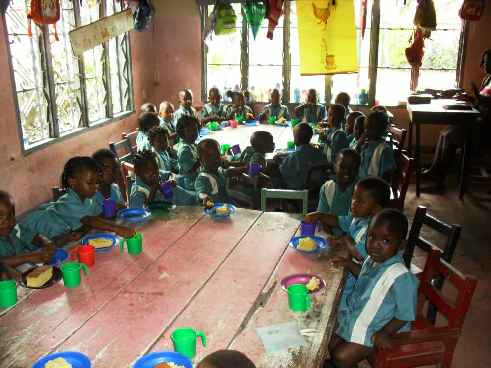 Young children in school uniform seated around long tables eating a hot meal.